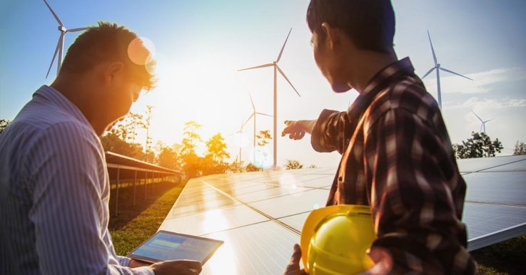 Two people looking out over a solar panel toward a wind farm as the sun rises.